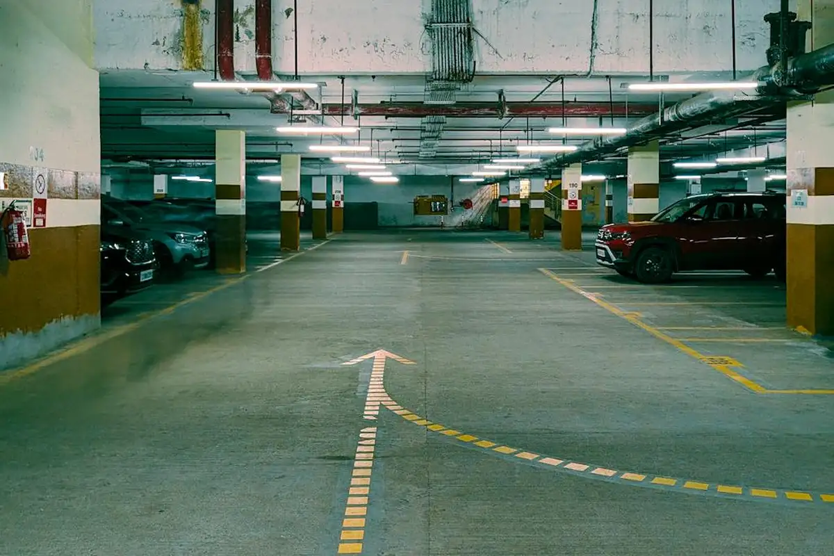 Indoor parking garage with concrete floor, yellow support columns, overhead pipes, and cars parked along the sides; a painted arrow guides traffic down the center.