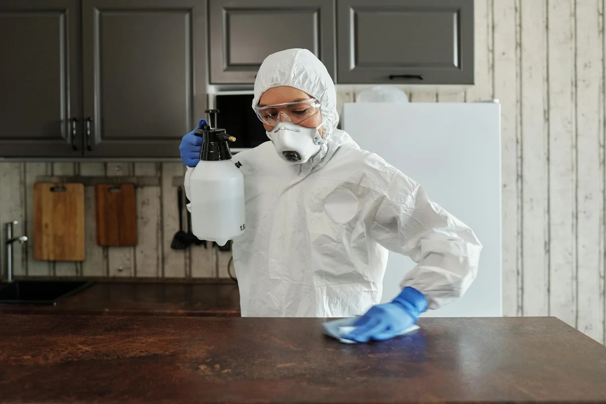Pest control worker wearing protective suit, gloves, and mask, spraying a countertop in a kitchen.