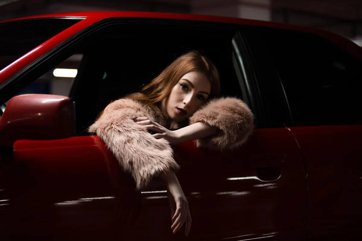 A person wearing a fur coat leans out of the window of a red car in a dimly lit setting.