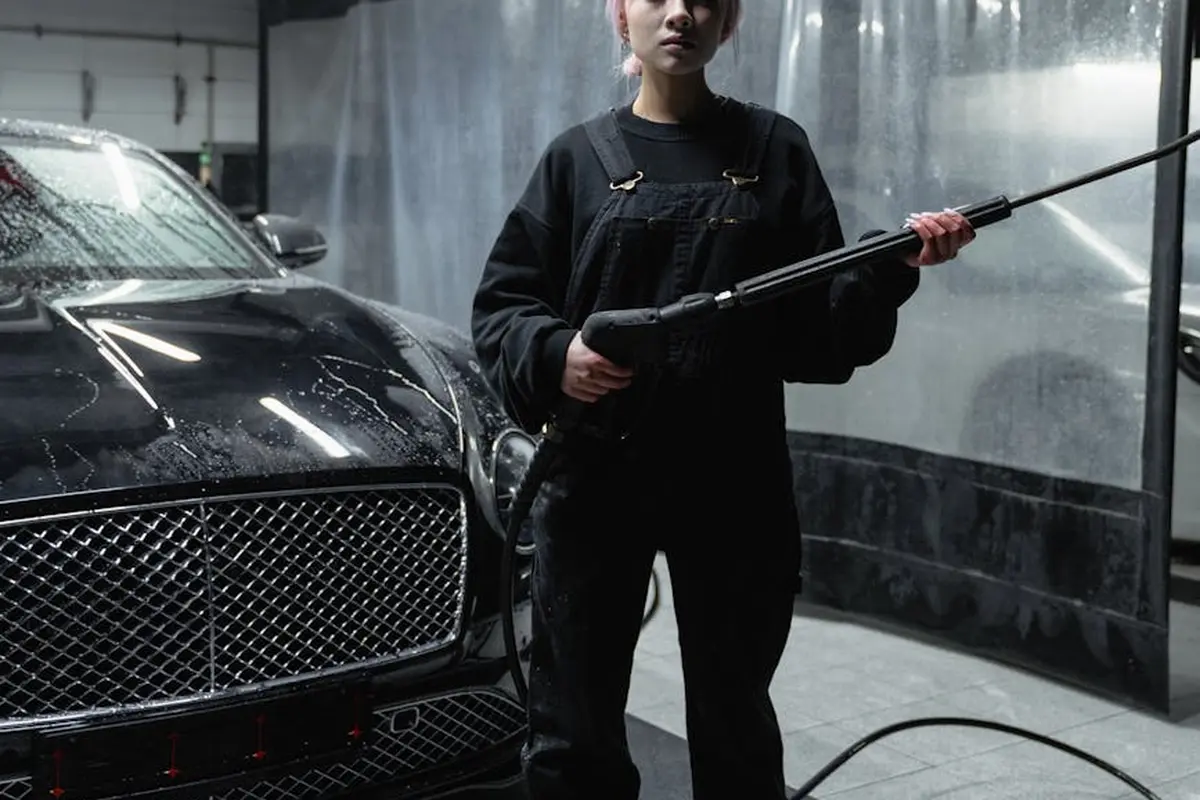 A person in dark overalls operates a pressure washer in a garage, preparing the concrete floor beside a black car.