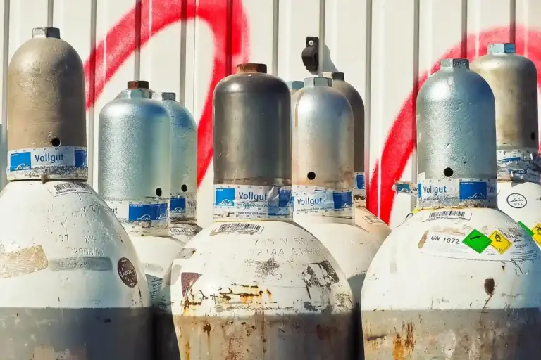 Row of propane gas cylinders with blue labels, some rust on the tanks, set against a wall with red graffiti.