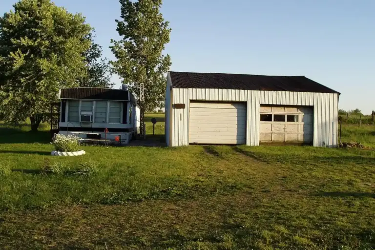 Rural property with a small mobile home beside a two-door metal garage on a grassy field with trees