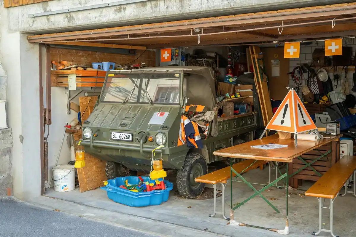 Open garage with a rugged military-style vehicle parked inside, surrounded by workbenches, tools, and orange safety signs.