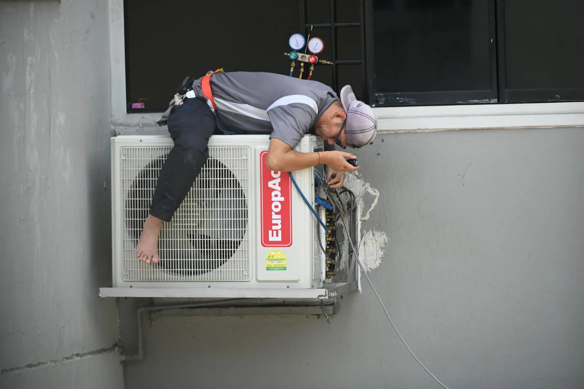 Technician crouched on an outdoor air conditioner condenser mounted to a garage wall, performing maintenance.