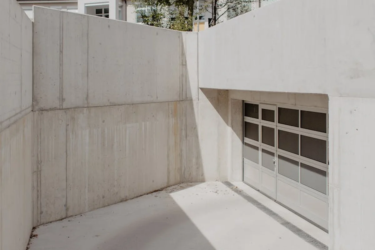 Concrete garage alcove with a closed sectional garage door; blank walls and sunlight casting a shadow, illustrating the space where a MyQ hub and sensor could be mounted.
