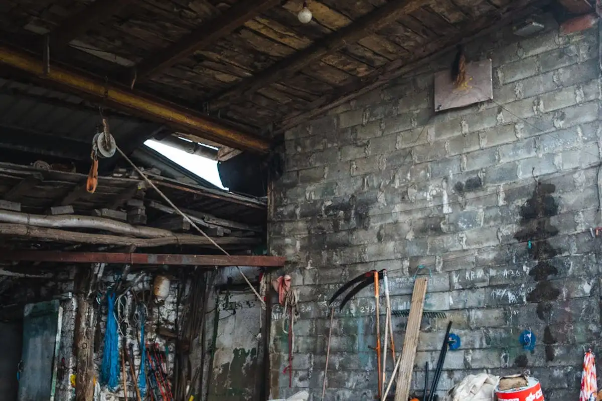 Cluttered garage interior with exposed ceiling beams, hanging tools, and a brick wall.