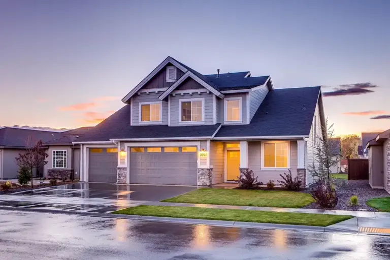Suburban two-car garage at dusk with a lit entry door, wet driveway, and manicured lawn.
