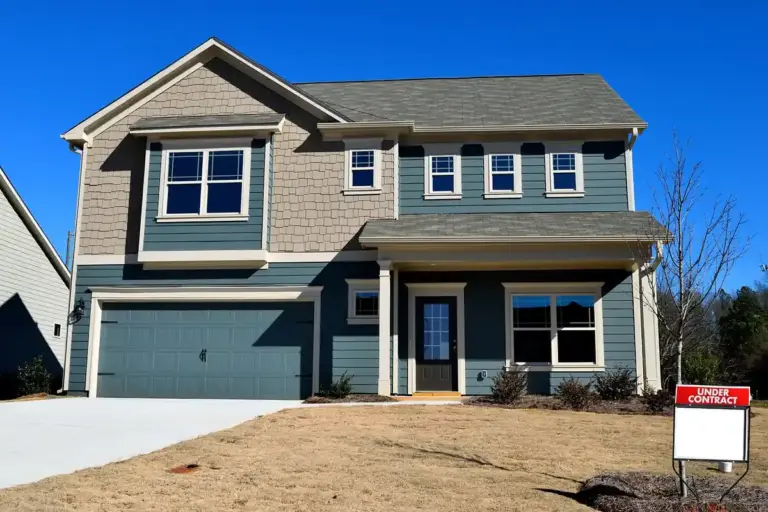 Front view of a two-story suburban home with a two-car attached garage and an 'Under Contract' sign in the front yard under a clear blue sky.
