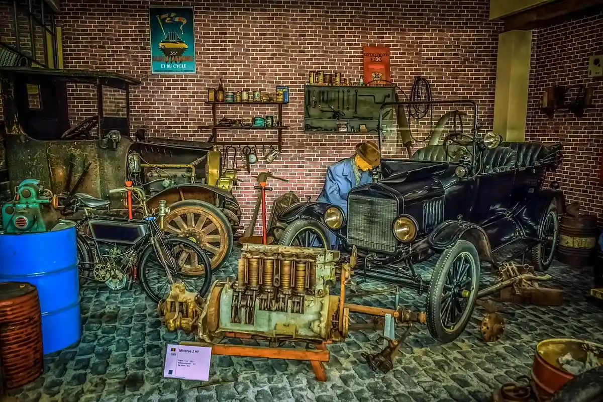 A vintage car garage with two old automobiles, tools on the wall, and a person working near one car on a cobblestone floor.