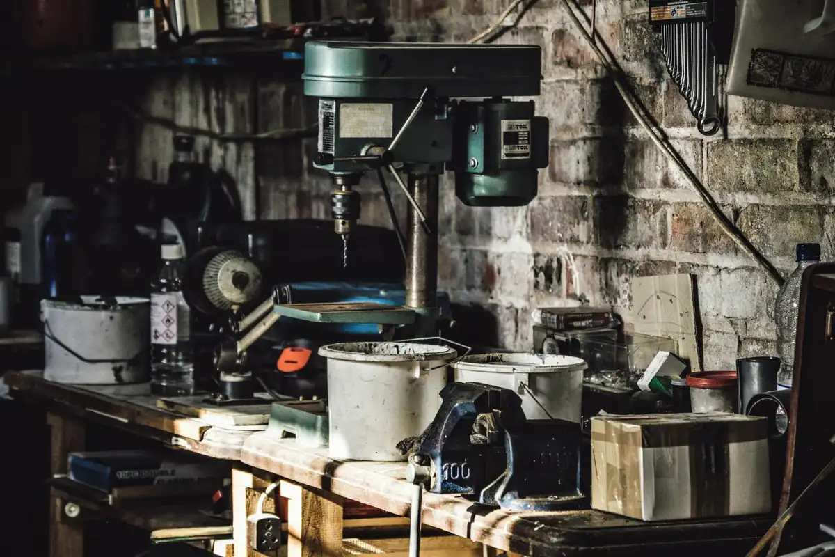 A dimly lit garage workshop with a drill press, tools, and containers spread across a wooden workbench.