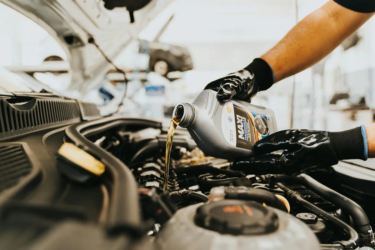 Mechanic wearing gloves pours motor oil into a car engine under an open hood.