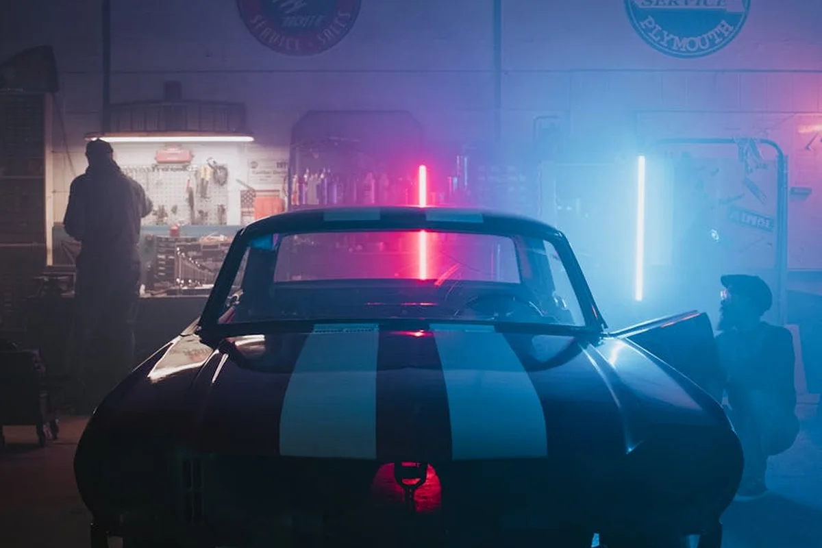 Rear view of a vintage blue racing car with white stripes inside a dim garage illuminated by red and blue neon lights, implying a restoration project that may require professional climate control.
