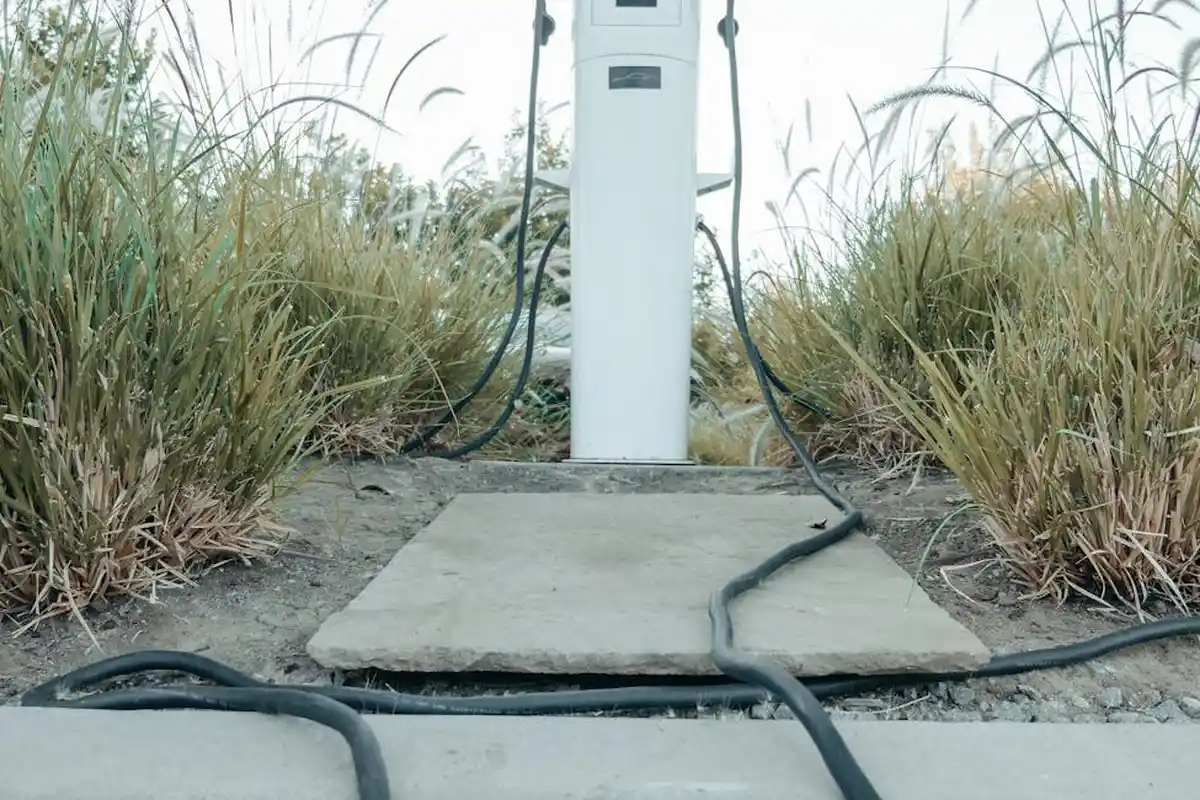 An electric vehicle charging pedestal mounted on a concrete pad outdoors, with thick cables running along the ground among tall grasses.