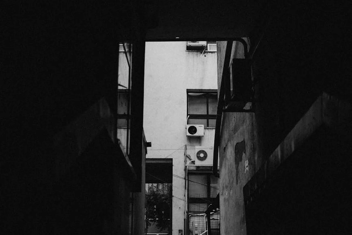 Black-and-white alley scene between tall buildings with exterior HVAC units mounted on the wall