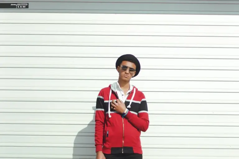 Person wearing a red, white, and black striped jacket, sunglasses, and a black hat standing in front of a white garage door.