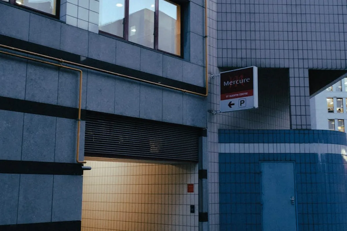 Exterior of a building with a partially open rolling garage door, illuminated interior, and a blue-gray facade; a Mercure hotel sign and parking symbol are visible.