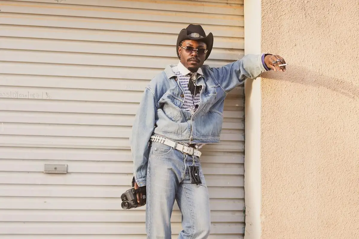 Man in denim jacket and jeans, wearing a cowboy hat and sunglasses, leaning against a beige wall beside a closed garage door, holding a camera