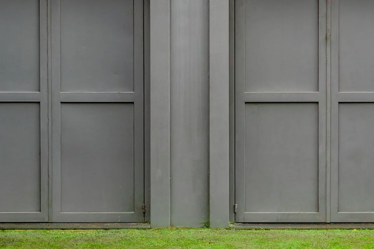 Two large gray garage doors with a narrow vertical seam in the middle and a strip of grass in front.