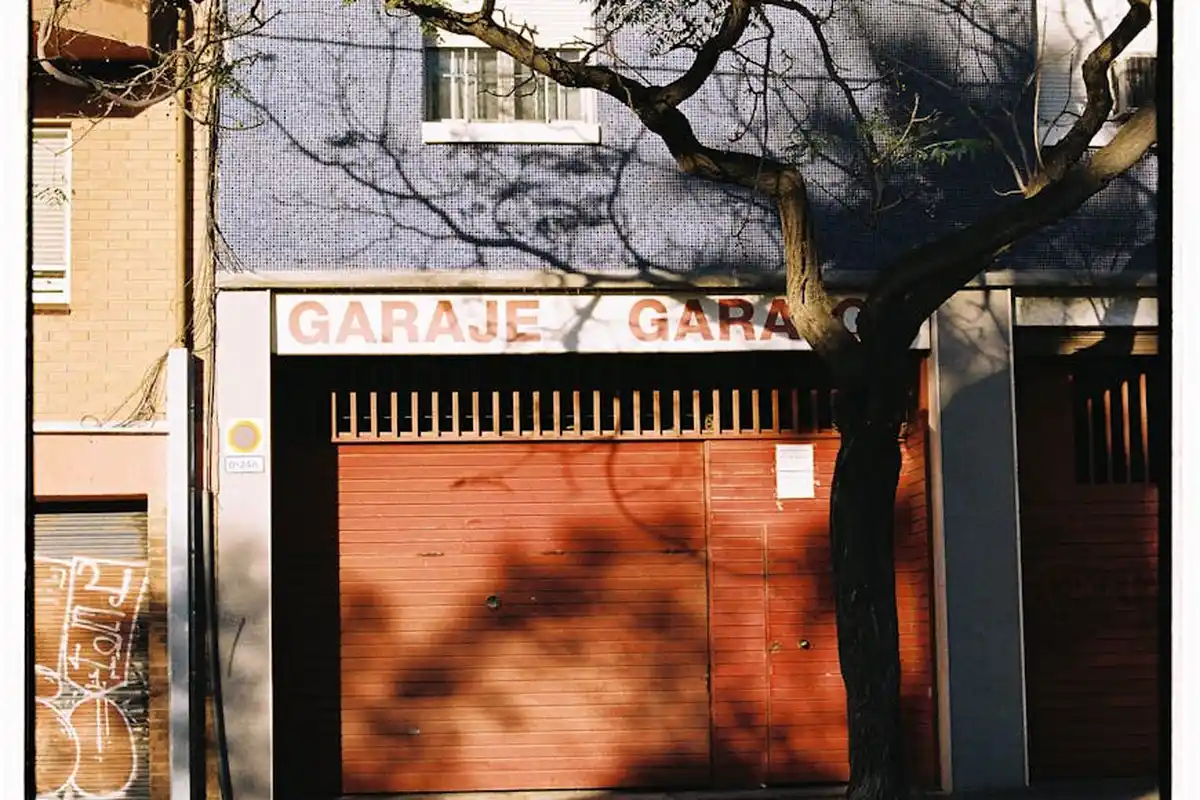 Residential garage with a closed red metal door, a tree in front, and a building façade.