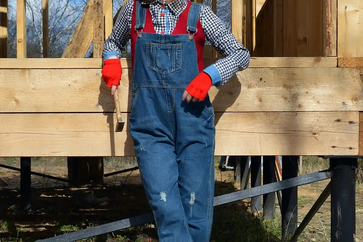 Construction worker in blue denim overalls and red gloves stands beside a partially framed wooden garage on raised supports.