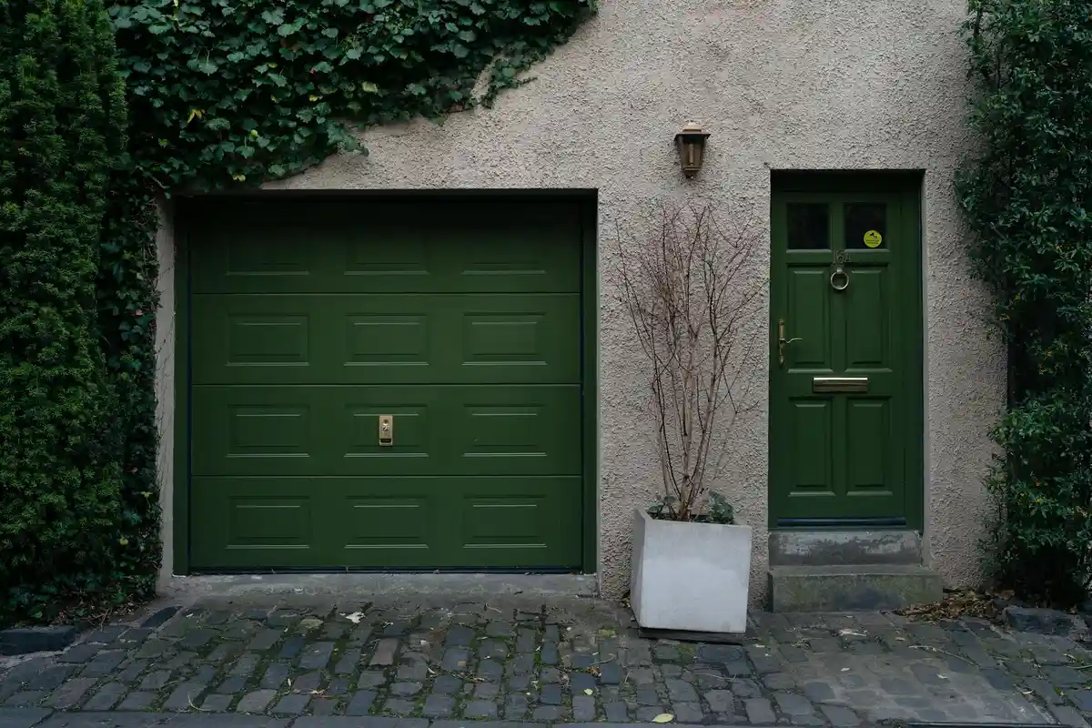 Green garage door with a matching entry door, a potted plant between doors, and ivy on the surrounding wall, set on a cobblestone driveway.