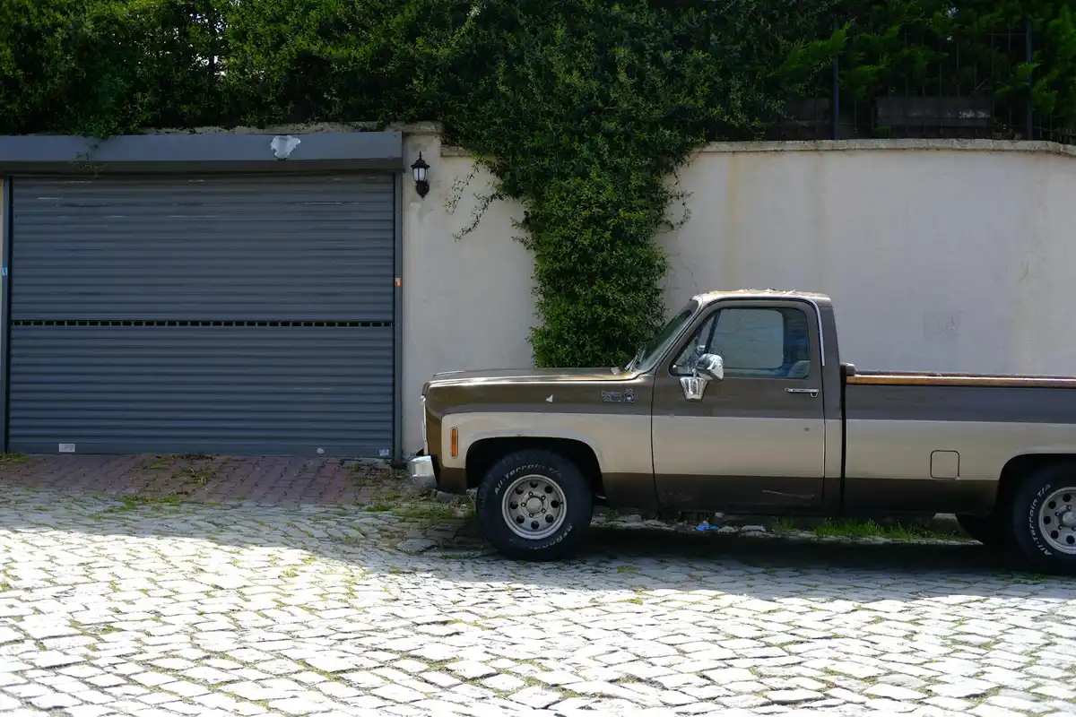 Garage exterior with a large roll-up door, white wall, greenery climbing a section of the wall, and a vintage pickup truck parked on a cobblestone driveway.