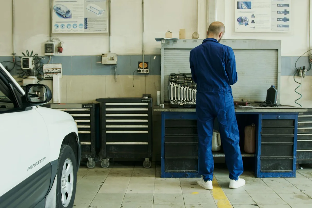 A technician in blue coveralls stands at a workbench in a garage workshop, with tool chests, equipment, and a car nearby.