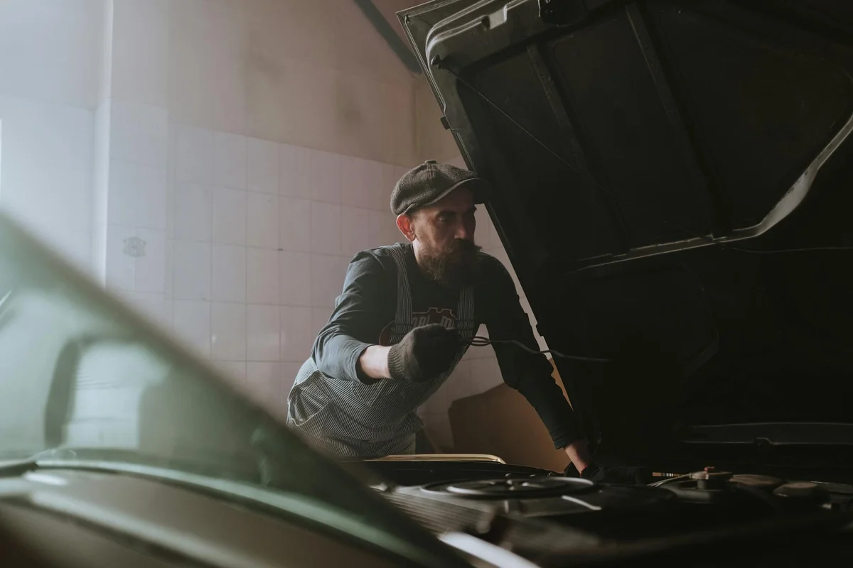 Mechanic wearing a cap and overalls works under the hood of a car in a garage workshop.