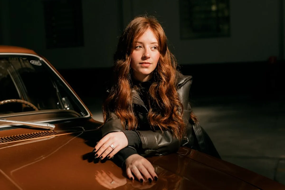 A person leaning on a vintage orange car inside a dimly lit garage.