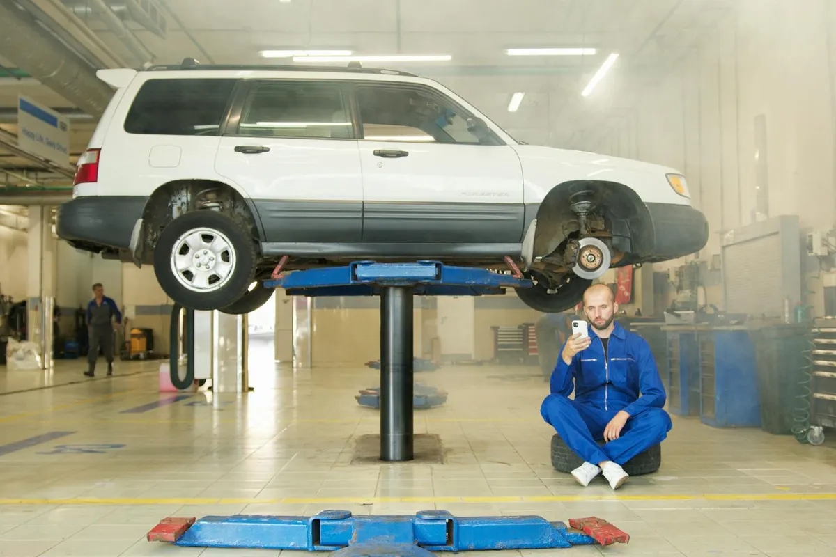 A mechanic in blue coveralls sits on the floor next to a white SUV raised on a hydraulic lift inside a well-lit garage, with tools and equipment visible in the background.