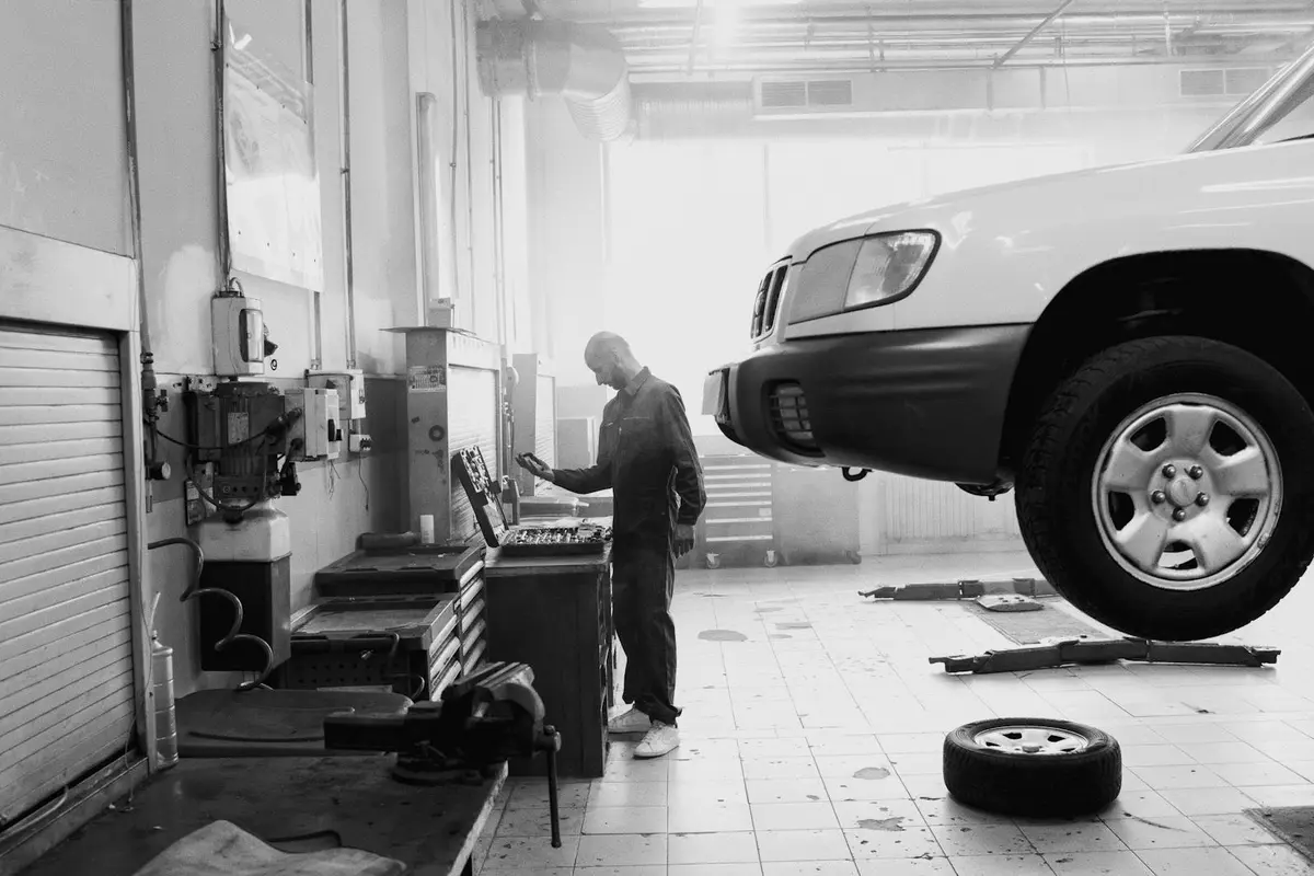 A mechanic stands beside an elevated vehicle in a bright workshop, using a laptop or diagnostic device at a workstation while tools and a spare tire lie nearby.