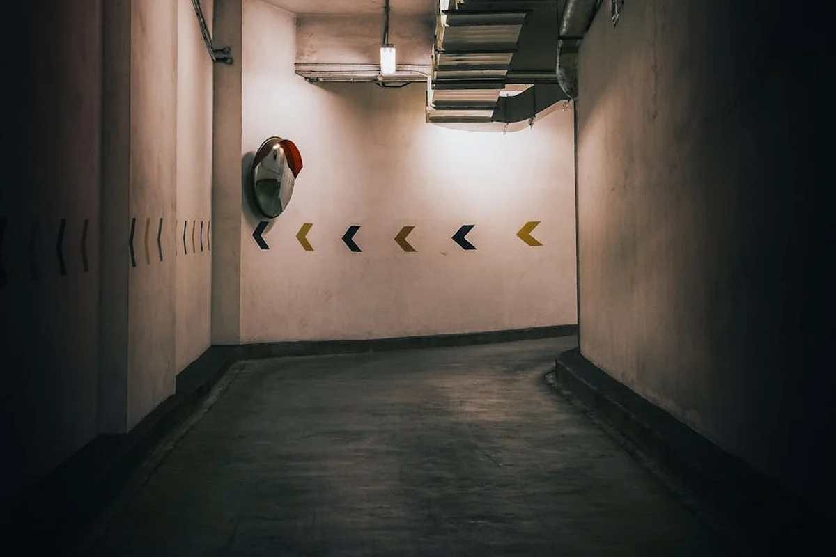 Dim underground parking garage with a narrow concrete corridor, yellow chevron arrows on the far wall, and a red safety device mounted on the left wall.