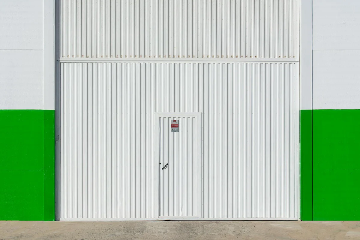White sectional garage door with a small pedestrian door in the center, flanked by green wall panels and a concrete driveway.