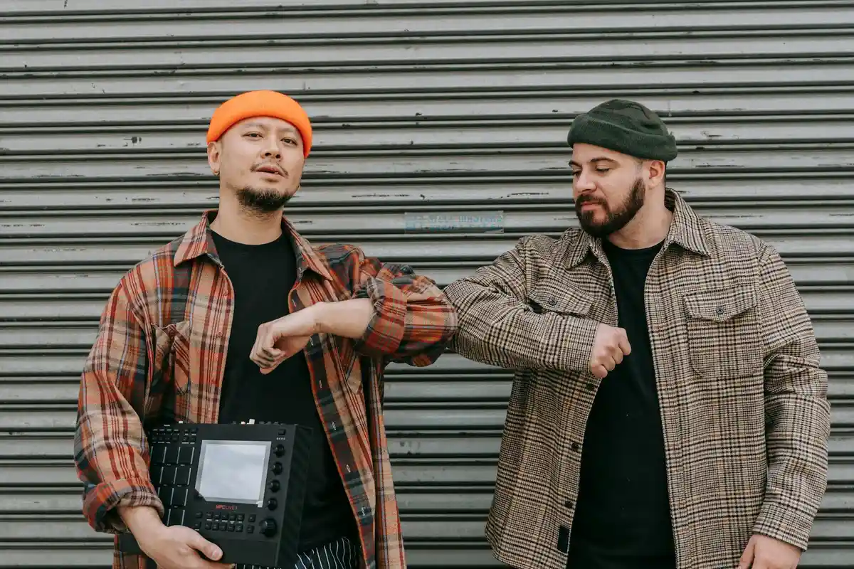 Two men in flannel shirts and beanies stand by a metal garage door; one holds a control panel while the other gestures with a raised fist, illustrating hands-on DIY work.