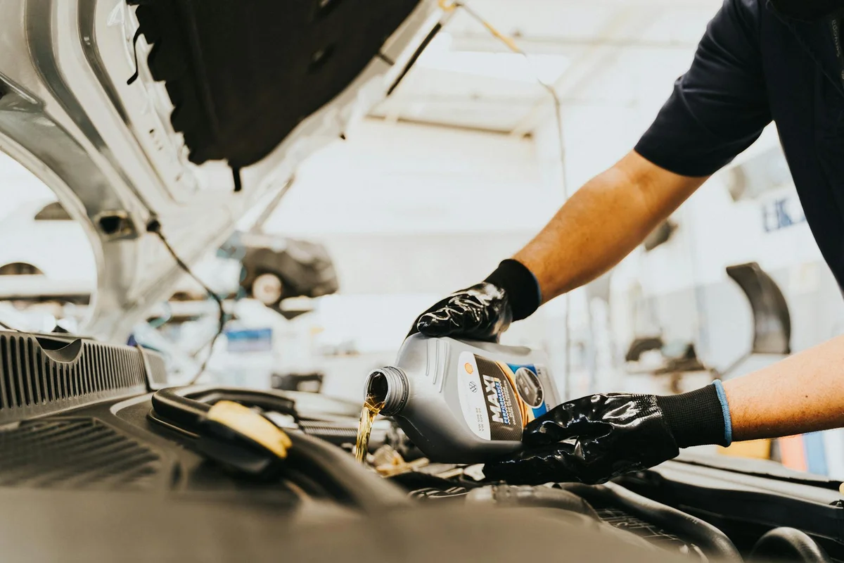 A mechanic wearing gloves pours engine oil into a car engine with the hood open in a bright workshop.