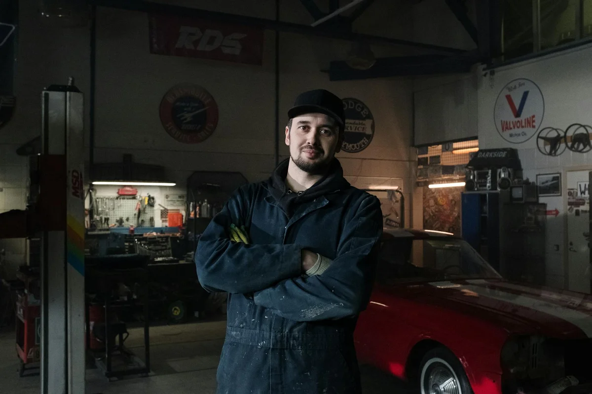 Portrait of a mechanic in blue coveralls and a cap standing with arms crossed inside a dimly lit garage beside a red car, representing a workshop environment.
