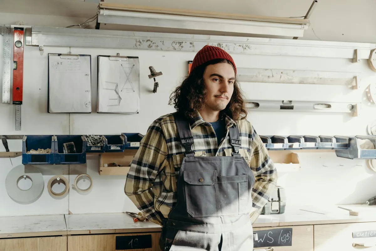 Woodworker in a garage workshop wearing a red beanie and plaid shirt, standing with hands on hips in front of a workbench with tools and dust-collection equipment in the background.