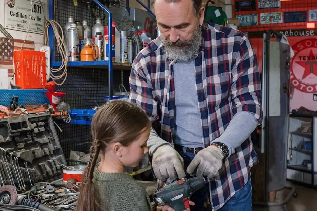 Bearded man in a plaid shirt and a young girl work together with a drill in a cluttered garage workshop.