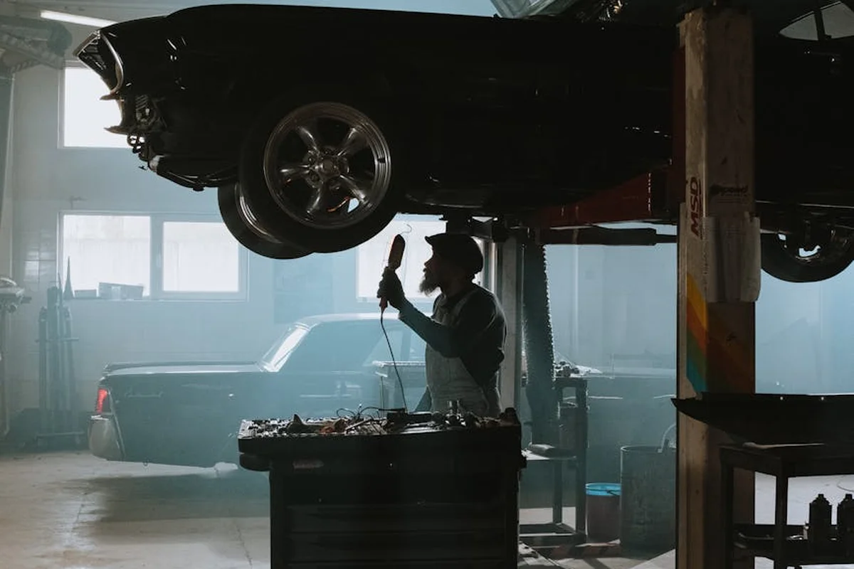 A person is working under a vehicle raised on a hydraulic lift in a cluttered workshop, with tool benches and another car visible in the background.