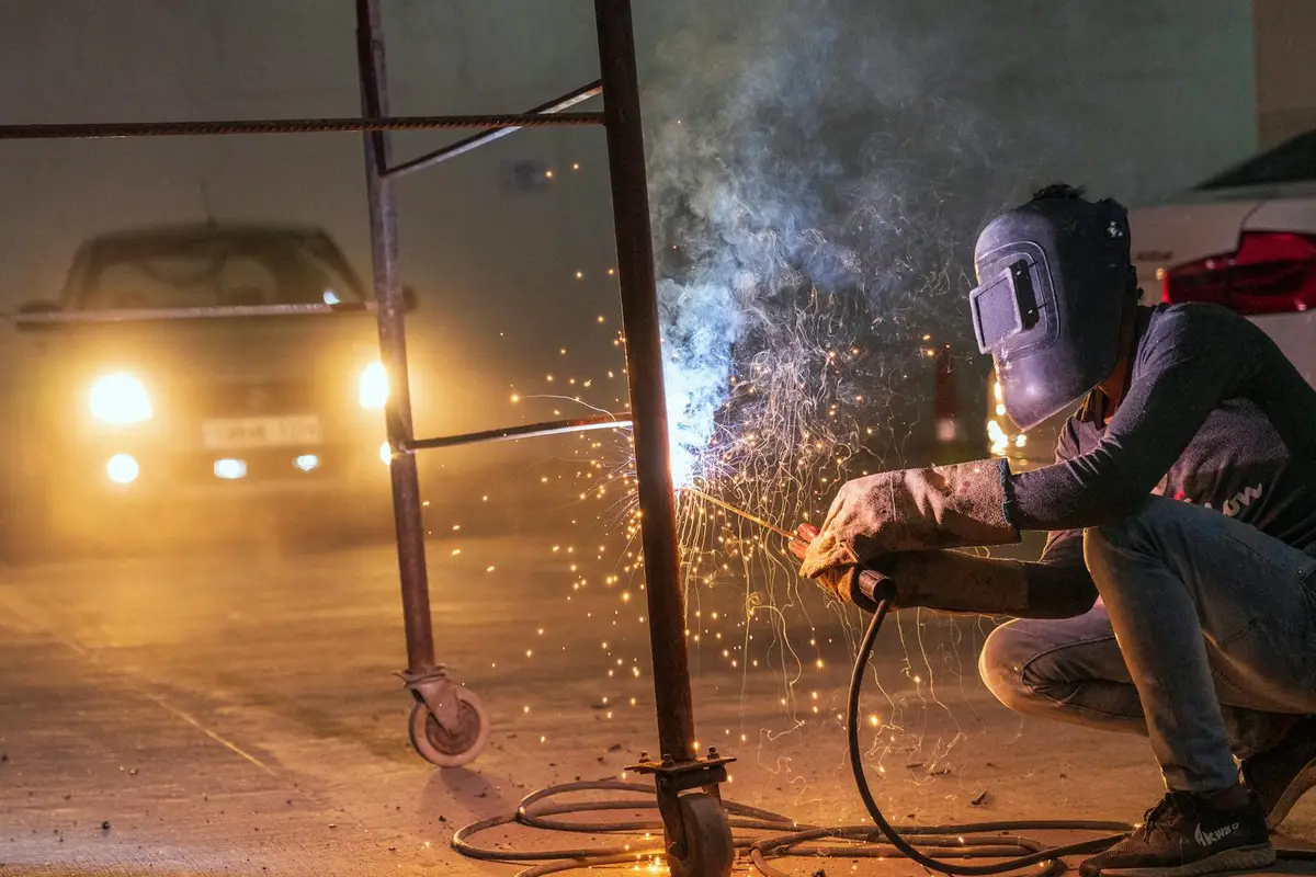 Welder in a garage workshop wearing a welding helmet crouches beside a metal frame, sparks flying as they weld, with car headlights glowing in the background.