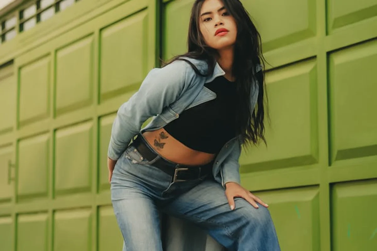 Woman with long dark hair wearing a denim jacket and jeans poses in front of a lime-green panel garage door.