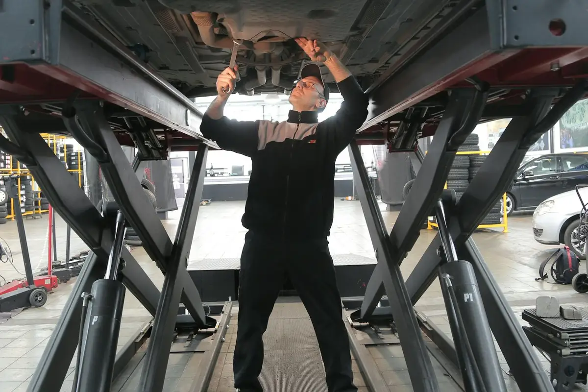 Technician crouched beneath a car on a hydraulic lift in a busy garage, examining components with tools in hand.