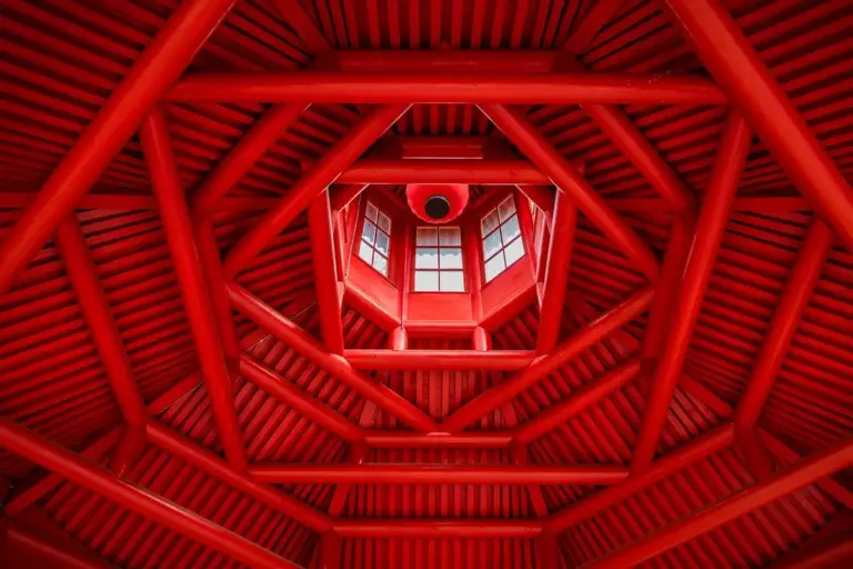 Interior view of red wooden roof trusses forming a geometric pattern around a central skylight.