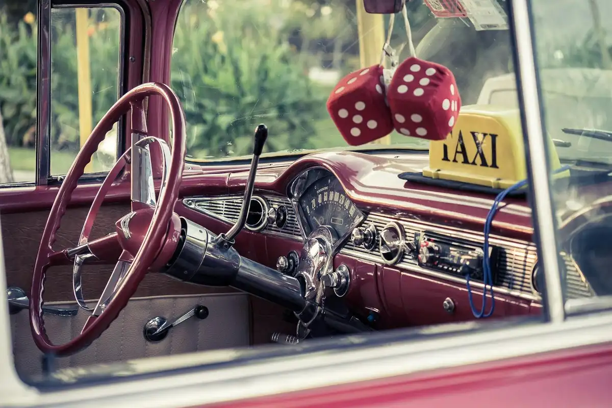 Red vintage car interior with a classic steering wheel, chrome dashboard, and hanging red dice, viewed through the window along with a taxi sign.