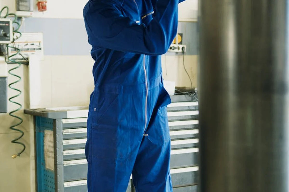 A person wearing blue coveralls standing in a workshop or garage with tool storage in the background